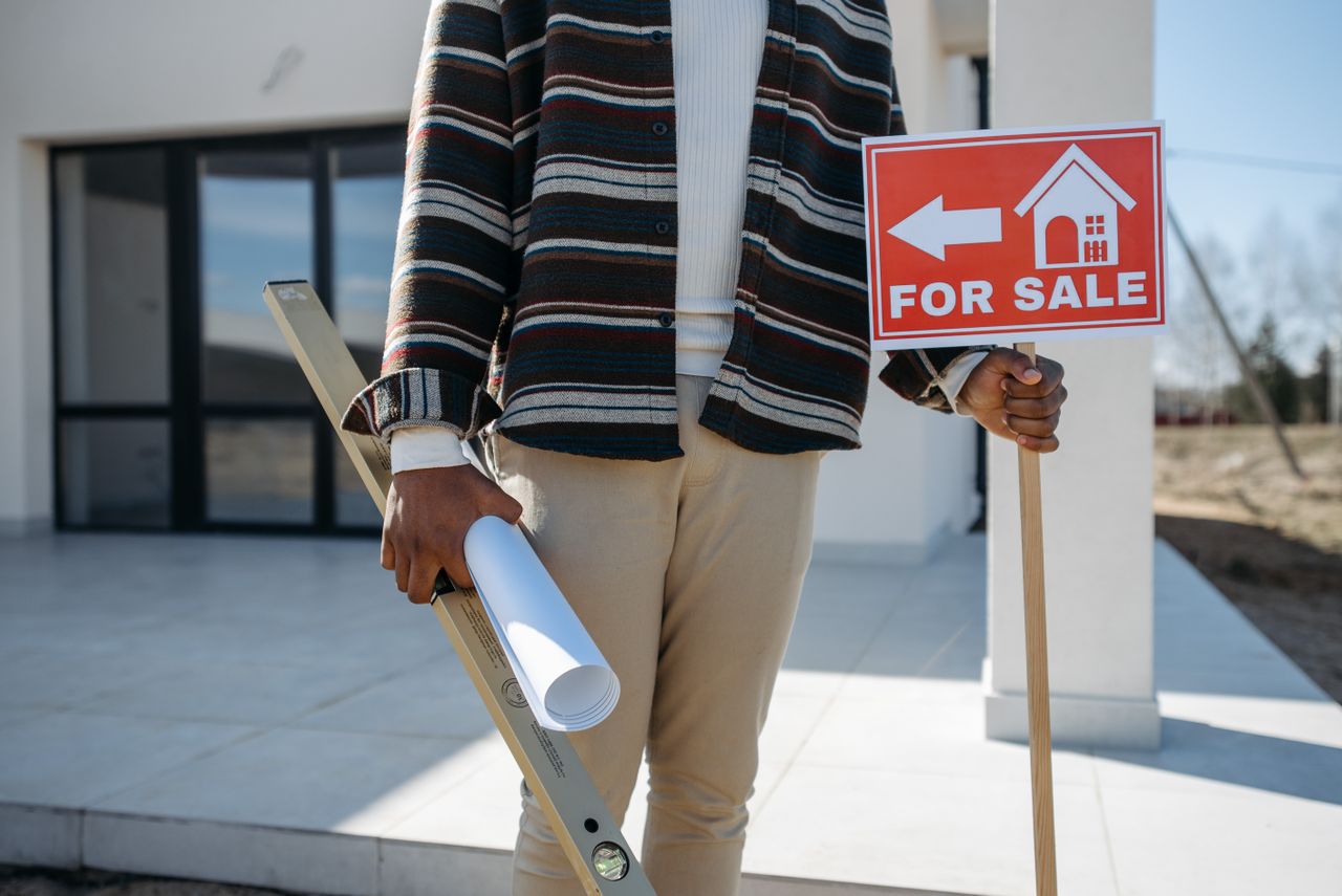 The Art of Drawing Readers In: Your attractive post title goes here A real estate agent stands outside with a 'for sale' sign and rolled blueprint, suggesting a property for sale.